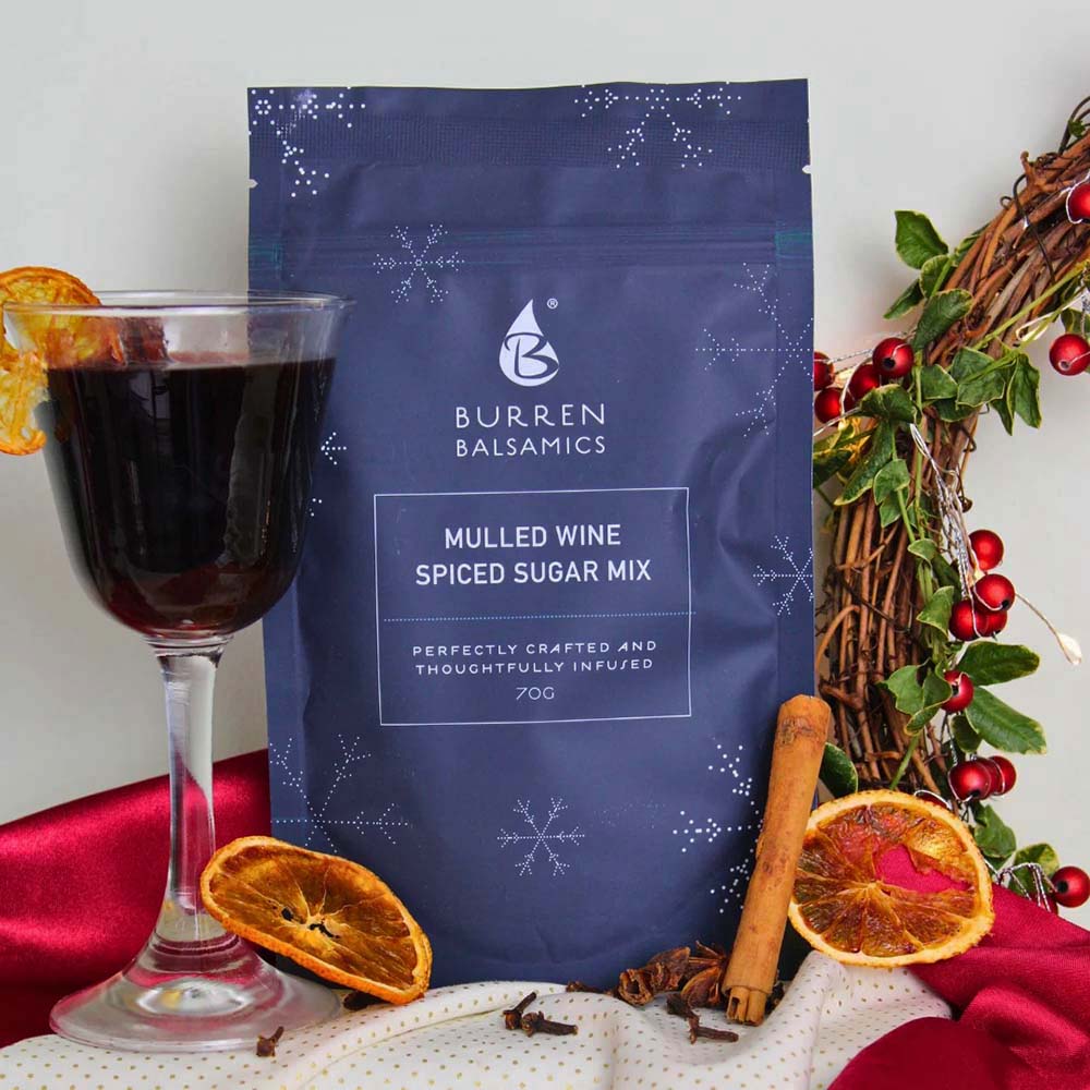 Festive holiday display featuring a pouch of Burren Balsamics Mulled Wine Spiced Sugar Mix alongside a glass of warm mulled wine, dried orange slices, cinnamon sticks, whole cloves, and a Christmas wreath.