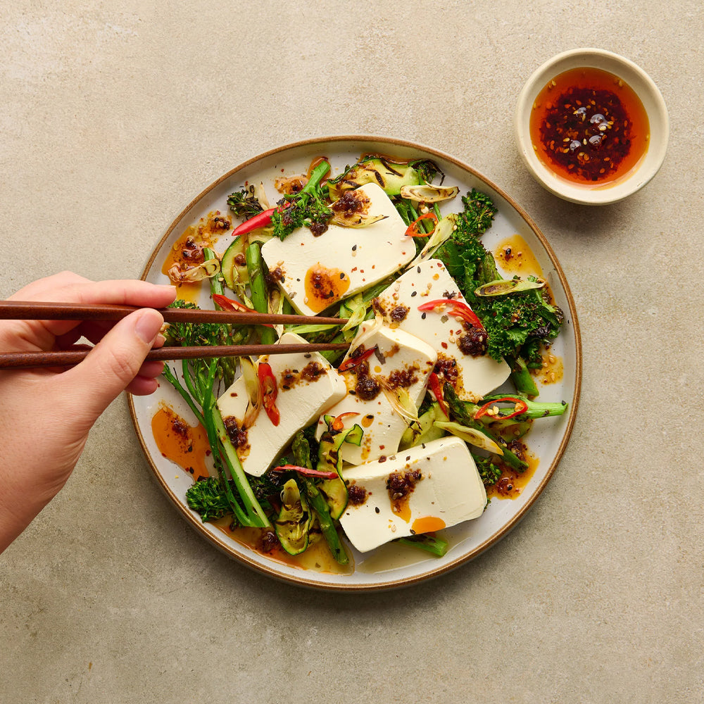 Plated dish of tofu and vegetables with a hand using chopsticks, on a neutral background.