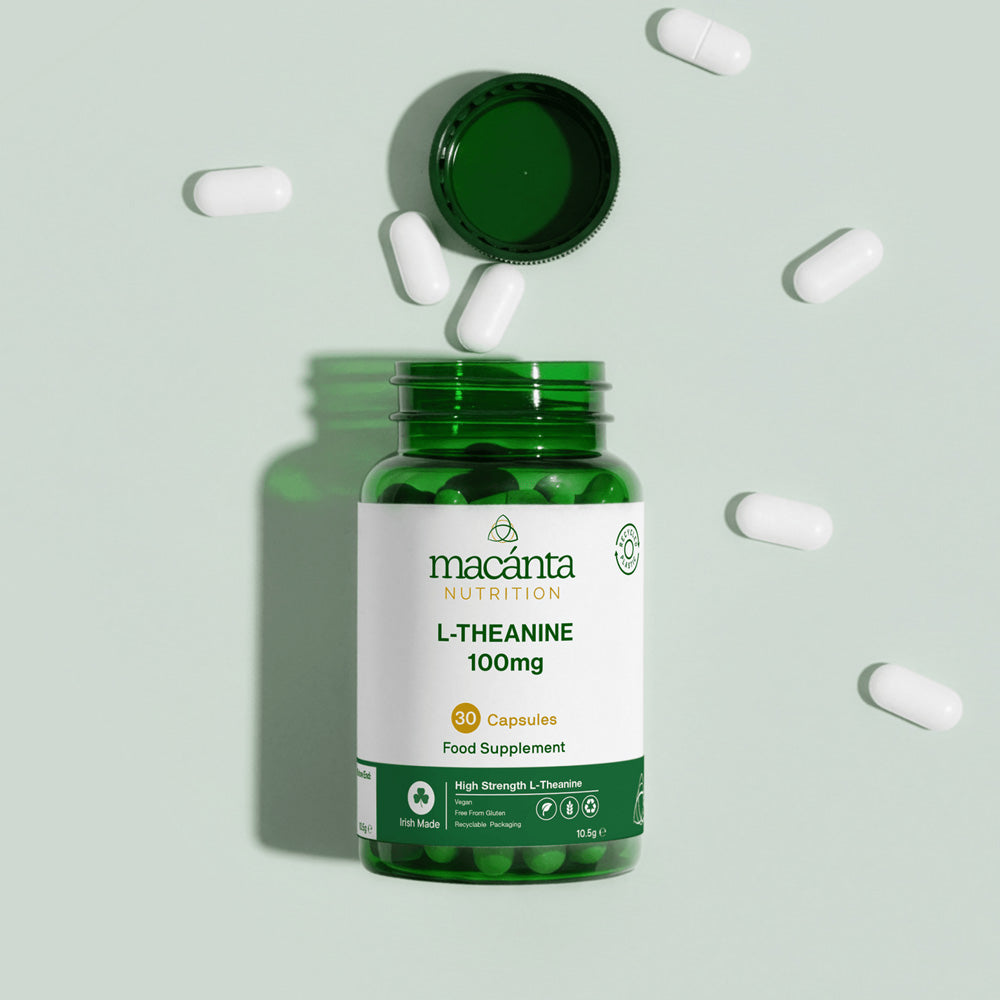 A green bottle of Macanta Nutrition L-Theanine 100mg capsules with the cap off, surrounded by scattered white capsules on a pale green background.