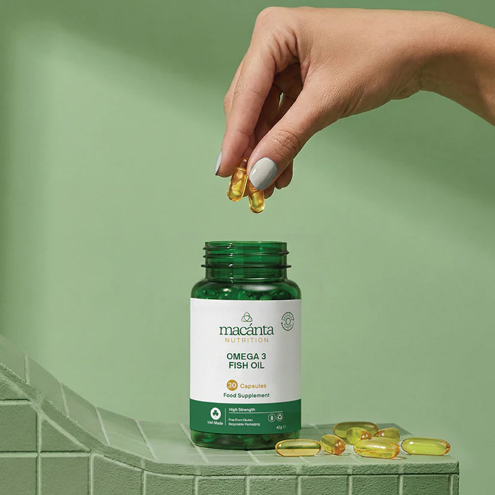 Hand holding fish oil capsules with a macánta Omega 3 Fish Oil bottle on a green background