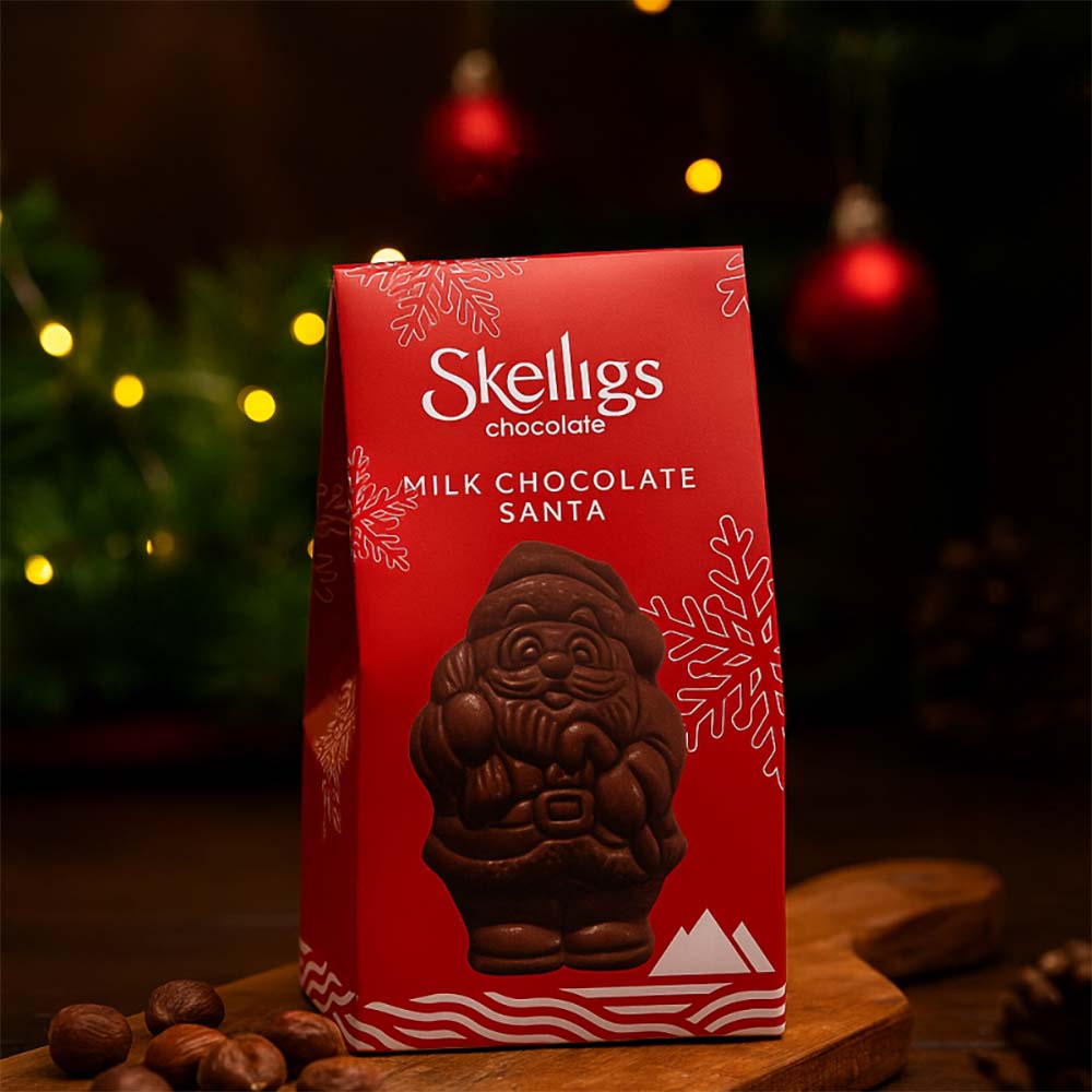 Festive holiday display of a Skelligs Chocolate Milk Chocolate Santa red gift box sitting on a wooden board, set against a warm, dimly lit background with blurred Christmas lights and ornaments.