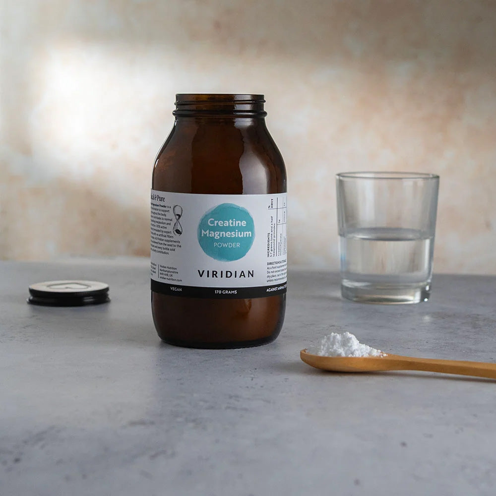 An amber glass jar of Viridian Creatine Magnesium supplement with its lid off, placed on a gray surface next to a glass of water and a wooden spoon with white powder.