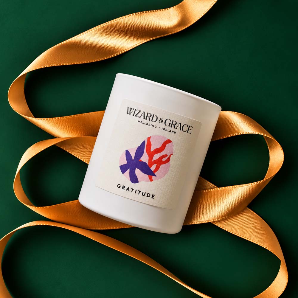 An overhead shot of the Wizard & Grace Gratitude candle in a white matte jar, framed by a swirling metallic gold satin ribbon against a deep forest green surface. The label reads "Wizard & Grace Wellbeing Ireland" and "Gratitude," featuring a colorful abstract design in purple, red, and pink.