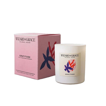 A white glass vessel candle and pink box for the Wizard & Grace "Gratitude" natural essential oil candle, featuring dark blue and red floral artwork.