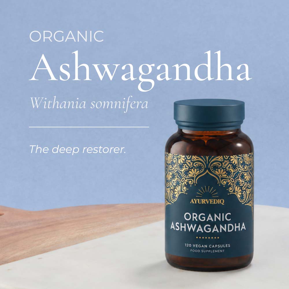 A bottle of Ayurvedic Organic Ashwagandha capsules with a dark blue label and gold ornamental pattern, set on a white surface with a wooden board. The background is a soft light blue with white text reading &
