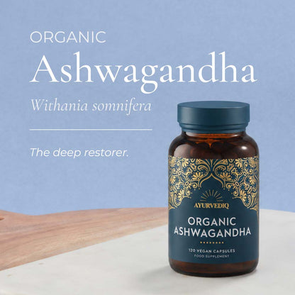 A bottle of Ayurvedic Organic Ashwagandha capsules with a dark blue label and gold ornamental pattern, set on a white surface with a wooden board. The background is a soft light blue with white text reading &