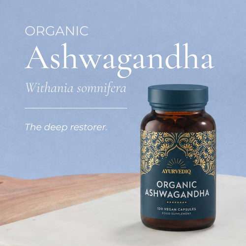 A bottle of Ayurvedic Organic Ashwagandha capsules with a dark blue label and gold ornamental pattern, set on a white surface with a wooden board. The background is a soft light blue with white text reading &