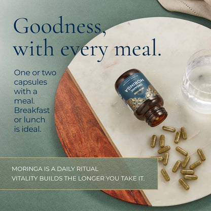 A bottle of Ayurvediq Moringa capsules lies on a  board with scattered capsules and a glass of water