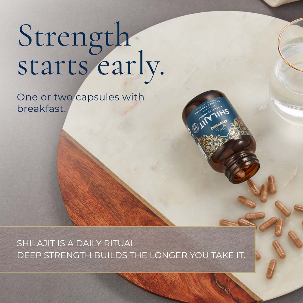 A bottle of Shilajit capsules lies on a white marble board with a wooden edge, surrounded by scattered brown capsules. A glass of water is partially visible at the top right, and promotional text appears on both sides.