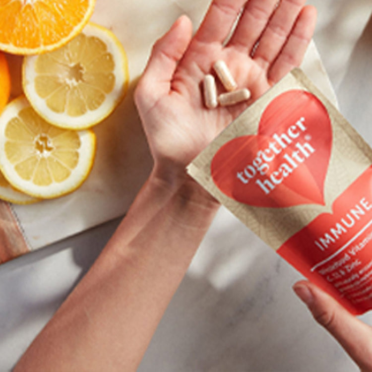 Hand holding immune supplement capsules with a 'Together Health' package and citrus fruits in the background.