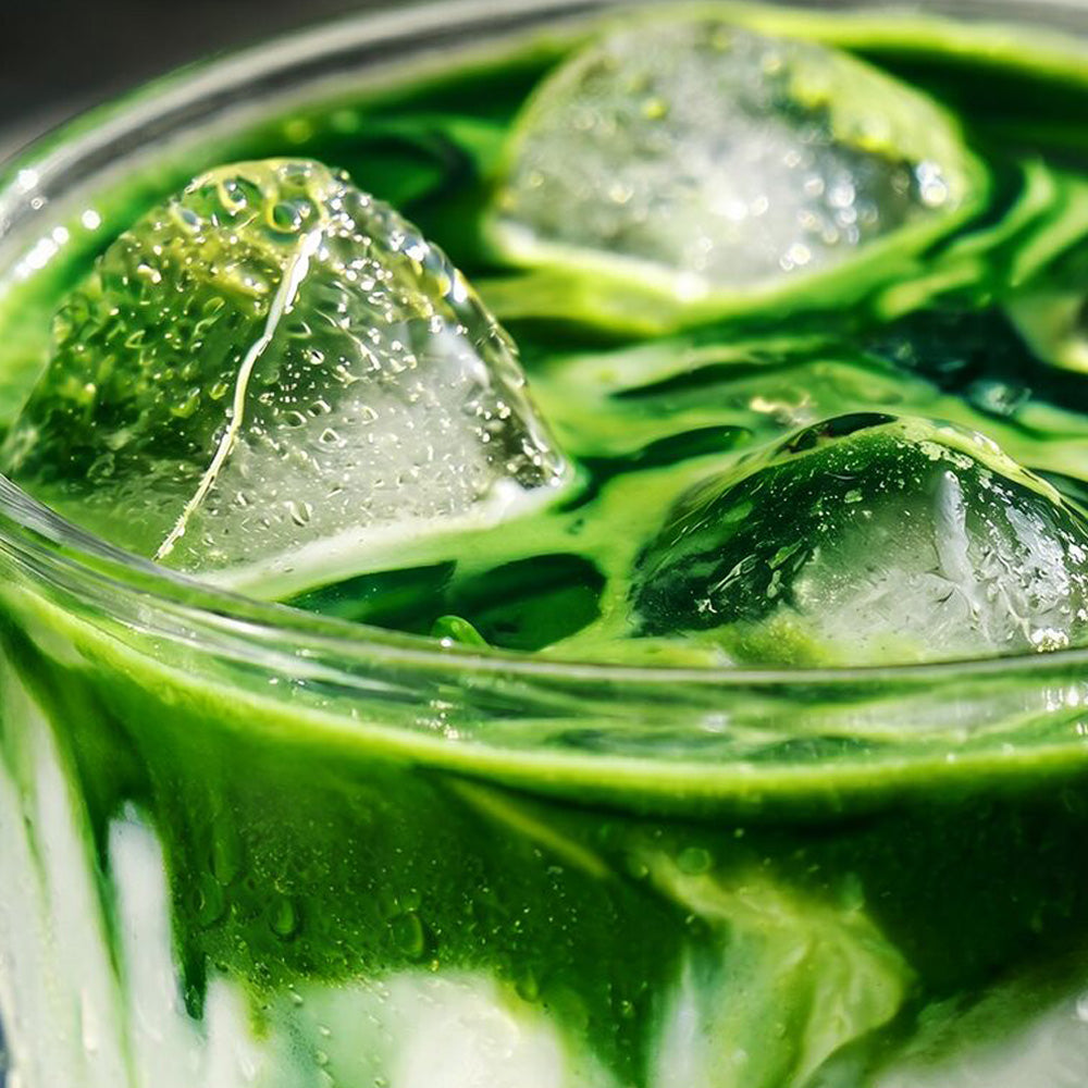 A close-up of a green iced drink in a clear glass, with large ice cubes and visible condensation droplets.