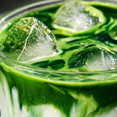 A close-up of a green iced drink in a clear glass, with large ice cubes and visible condensation droplets.
