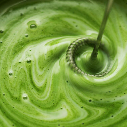 A close-up of a green frothy liquid being whisked in a circular motion, with visible bubbles and swirls in the texture.