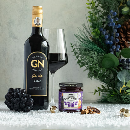 Bottle of Graham G. Norton Shiraz wine, a glass of wine, a jar of Ballymaloe Mulled Wine Cream Sauce, and grapes on a decorative surface with greenery.