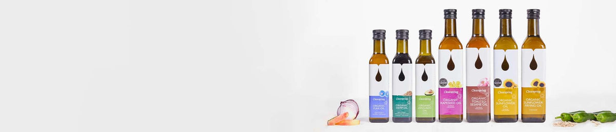 Six glass bottles of olive oil with black caps and colorful labels, arranged on a white background with small food props like garlic, onion, and parsley.