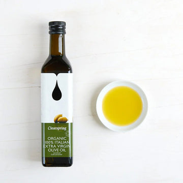 A bottle of Clearspring Organic 100% Italian Extra Virgin Olive Oil next to a small white bowl filled with the golden-yellow olive oil.