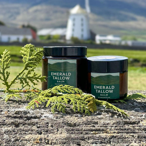 Two jars of Emerald Tallow Balm on a stone surface with a scenic background.