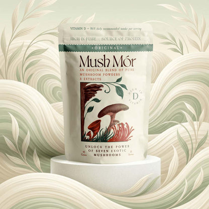 A stand-up pouch of Mush Mór mushroom powders and extracts centered on a white pedestal, with a decorative illustrated label showing mushrooms and botanical elements. The background is a soft, stylized cream and sage abstract floral pattern.