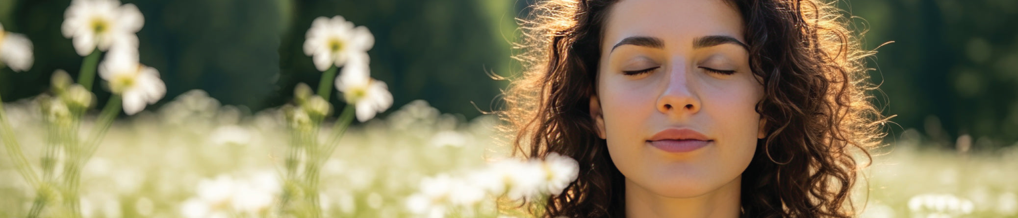 A close-up of a woman with curly brown hair standing outdoors in a sunlit field of white flowers, with her eyes closed and a serene expression.
