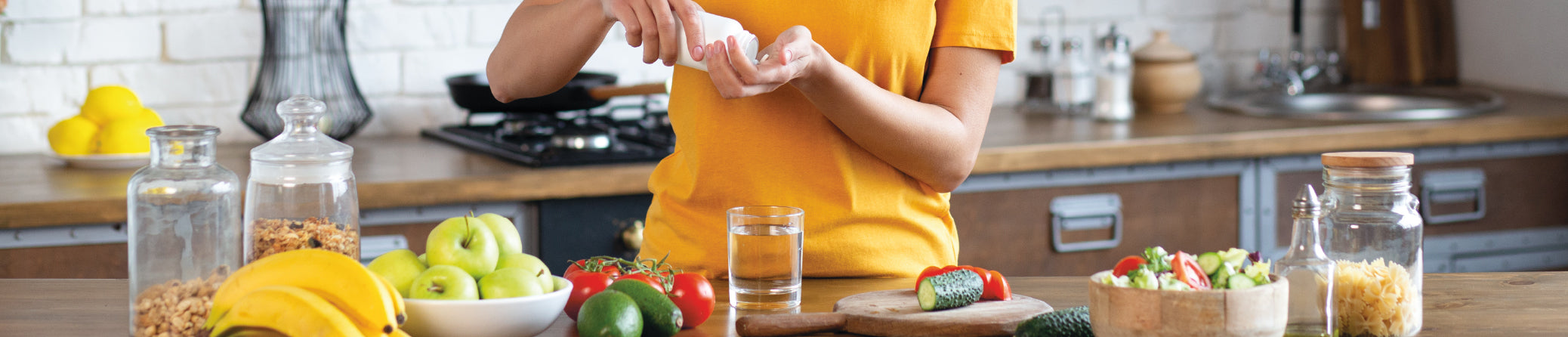 A person in a yellow shirt stands at a kitchen counter preparing food, with jars, fruits, vegetables, and a glass of water in a bright kitchen setting.