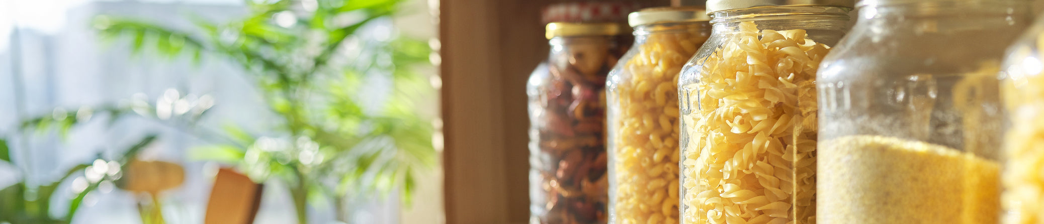 A close-up, slightly angled view of glass jars filled with dry pasta and other pantry items on a wooden shelf in a kitchen setting. The background is softly blurred with warm light and green foliage.