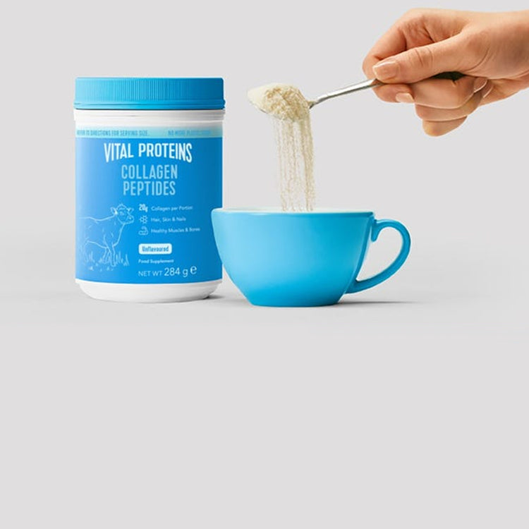 A hand holding a spoon over a blue cup, with a tub of vital proteins collagen powder beside it on a light gray background.