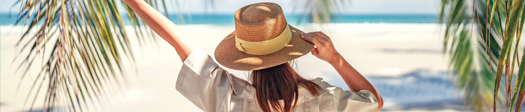A woman on a beach wearing a straw hat with a yellow band and a white shirt, with palm leaves framing the scene.