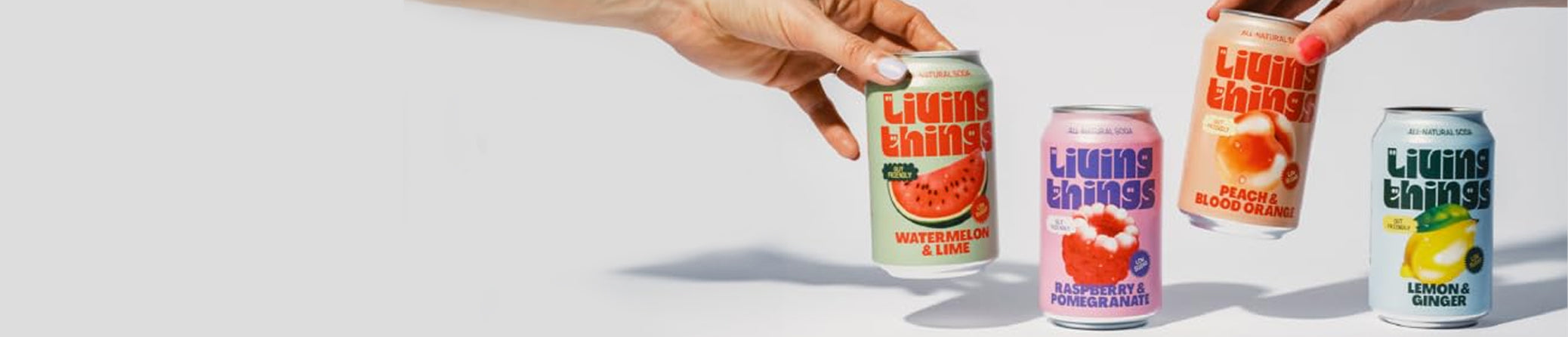 Two hands holding cans of Living Things Soda against a light gray background, with four colorful beverage cans lined up below them.