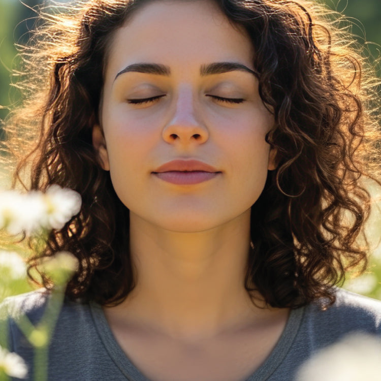 A close-up portrait of a woman with curly brown hair, eyes closed, wearing a gray t-shirt, set against a soft-focus outdoor background with warm sunlight.