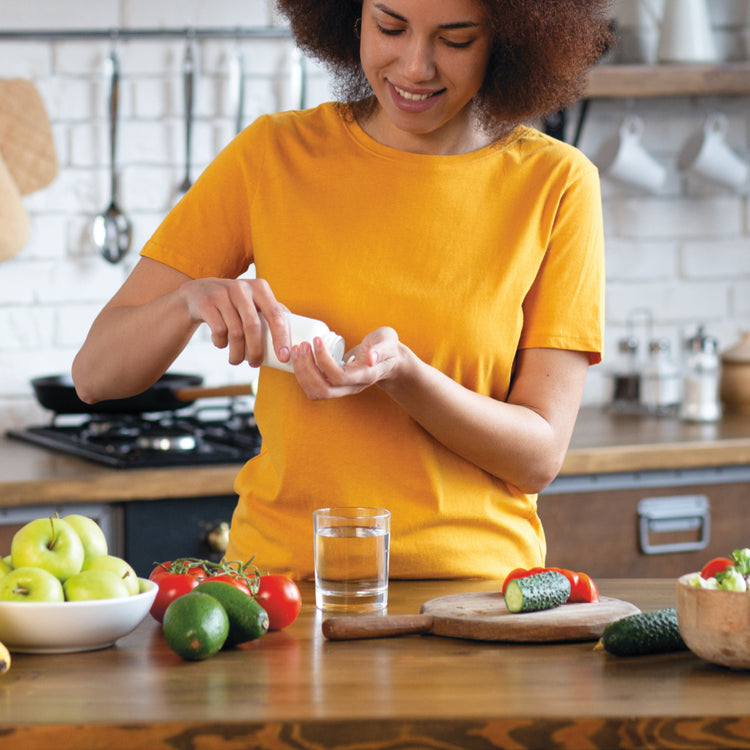A person in a yellow shirt stands at a kitchen counter preparing food, with jars, fruits, vegetables, and a glass of water in a bright kitchen setting.