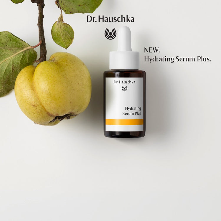 A small white dropper bottle labeled Dr. Hauschka Hydrating Serum Plus stands on a white background beside a green leafy fruit.