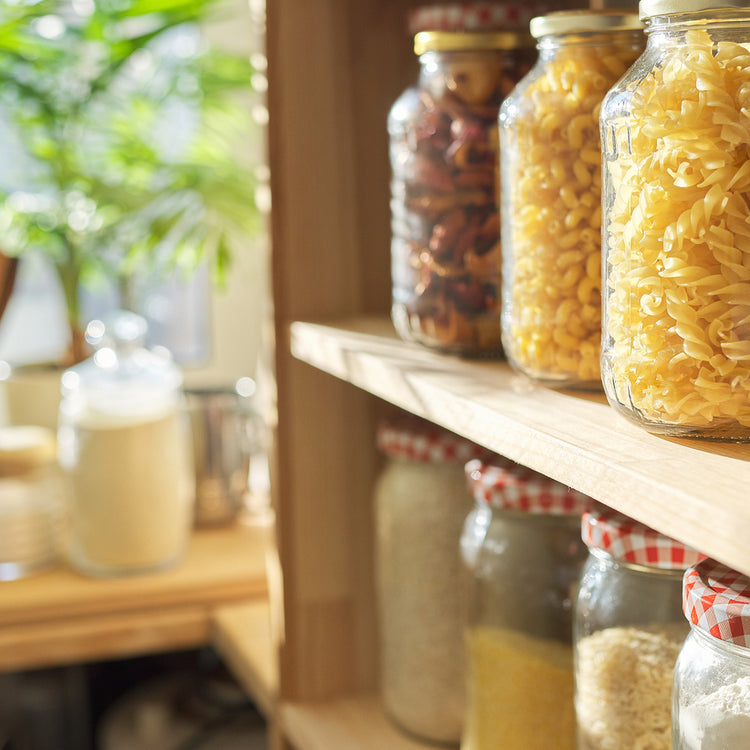 A close-up, slightly angled view of glass jars filled with dry pasta and other pantry items on a wooden shelf in a kitchen setting. The background is softly blurred with warm light and green foliage.