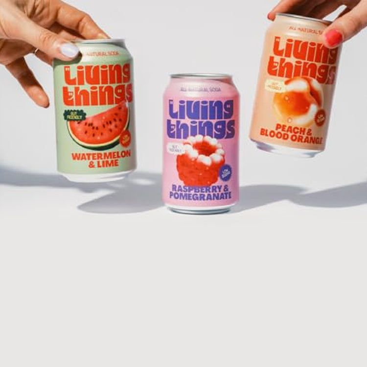 Two hands holding cans of Living Things Soda against a light gray background, with three colorful beverage cans lined up below them.