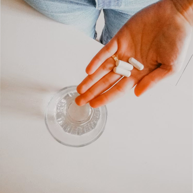 A close-up of a hand holding three white capsules above a clear glass, with a person's jeans visible at the top against a plain light background.