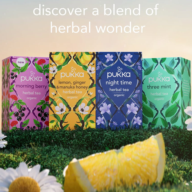 Four Pukka herbal tea boxes arranged in a grassy outdoor-style setting with a lemon slice and daisies in the foreground. 
