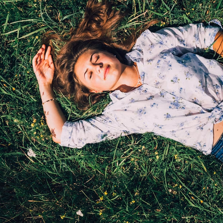 A woman sleeping, lying on her back in green grass, wearing a light blue patterned button-up shirt and brown arm sleeves.