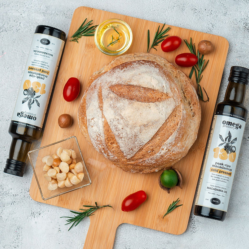 Loaf of bread on a wooden cutting board with olive oil bottles and Omega Pressery Cold Pressed Extra Virgin Macadamia Oil