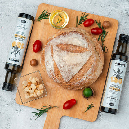 Loaf of bread on a wooden cutting board with olive oil bottles and Omega Pressery Cold Pressed Extra Virgin Macadamia Oil
