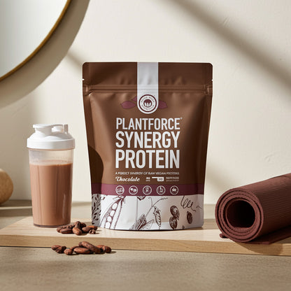 Plantforce Synergy Protein Chocolate flavour pouch displayed on a wooden board next to a shaker bottle with mixed drink, coffee beans, and a rolled yoga mat on a beige background.