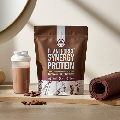 Plantforce Synergy Protein Chocolate flavour pouch displayed on a wooden board next to a shaker bottle with mixed drink, coffee beans, and a rolled yoga mat on a beige background.