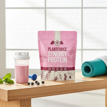 Plantforce Synergy Protein packet on a wooden surface with a pink shake, blue dumbbell, and green mat.