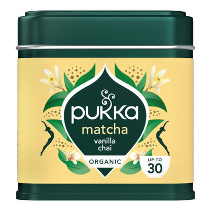 A tin of Pukka Matcha Vanilla Chai organic tea, featuring a dark green metal container with a pale yellow label decorated with green leaves, white flowers, and black silhouettes.