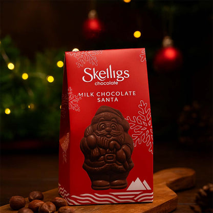 Festive holiday display of a Skelligs Chocolate Milk Chocolate Santa red gift box sitting on a wooden board, set against a warm, dimly lit background with blurred Christmas lights and ornaments.