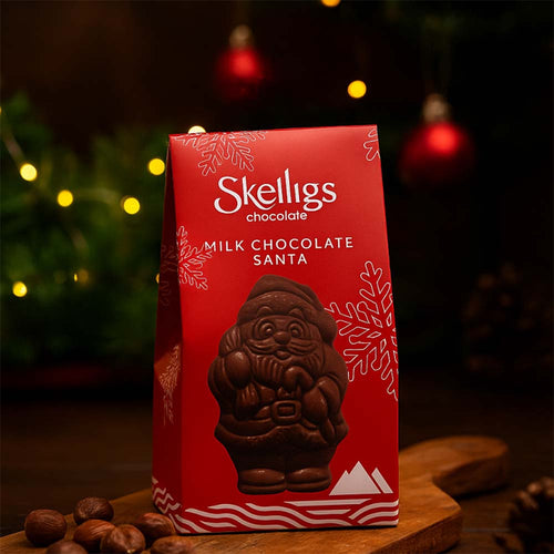 Festive holiday display of a Skelligs Chocolate Milk Chocolate Santa red gift box sitting on a wooden board, set against a warm, dimly lit background with blurred Christmas lights and ornaments.