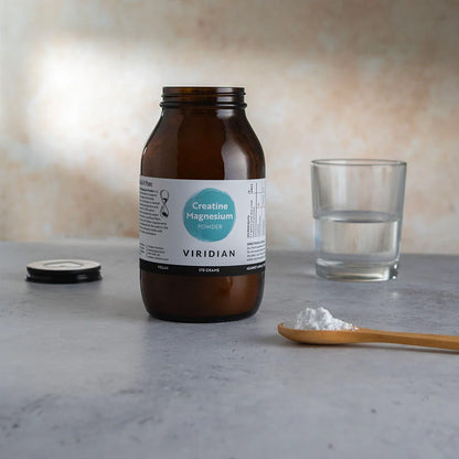 An amber glass jar of Viridian Creatine Magnesium supplement with its lid off, placed on a gray surface next to a glass of water and a wooden spoon with white powder.