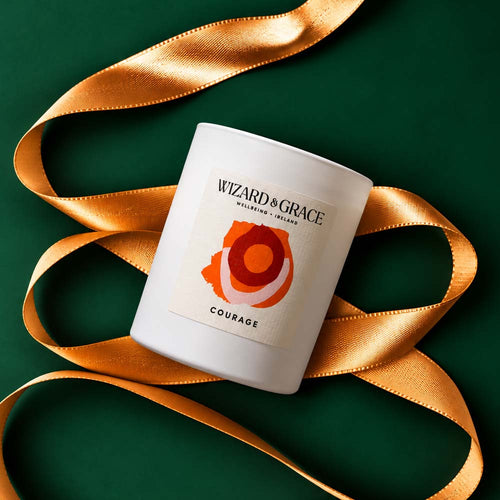 Top-down view of a white Wizard & Grace Courage candle resting on a dark green background surrounded by a shimmering gold ribbon.