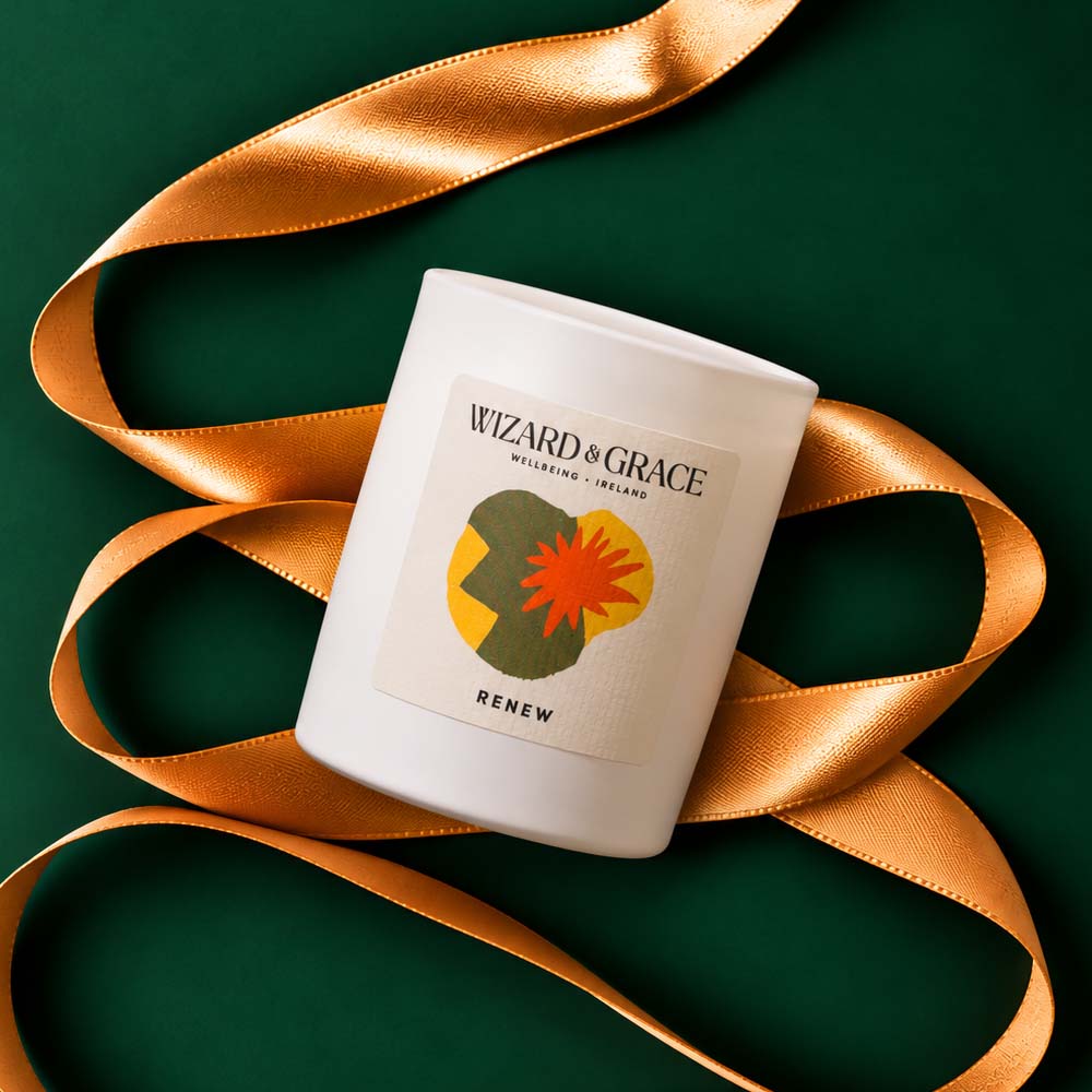 Top-down view of a white Wizard &amp; Grace "RENEW" candle jar resting on a dark green background, framed by a looping gold ribbon.