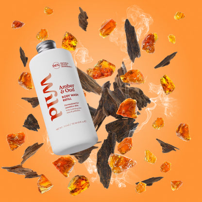 A bottle of Wild Amber &amp; Oud Scrub Wash floating over scattered amber pieces and dark wood shavings on an orange background.