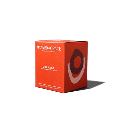 A bright orange square packaging box for the Wizard &amp; Grace Courage natural essential oil candle, featuring abstract layered oval artwork on the side.