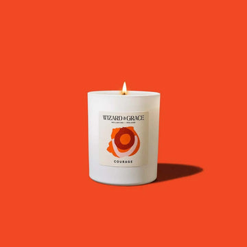A lit Wizard &amp; Grace Courage candle in a white glass vessel set against a solid bright orange background.